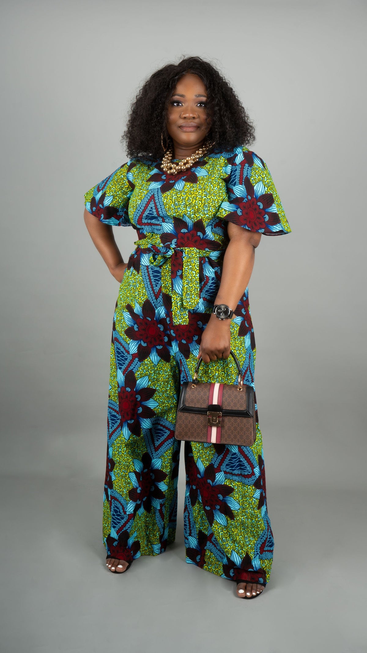 A woman modeling a vibrant green, blue, and red floral print wide-leg jumpsuit, holding a brown monogram handbag against a solid gray studio background at Bahati Design.