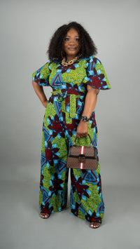 A woman modeling a vibrant green, blue, and red floral print wide-leg jumpsuit, holding a brown monogram handbag against a solid gray studio background at Bahati Design.