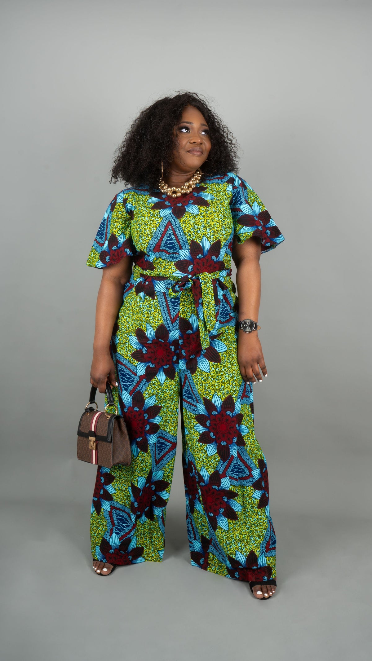 A woman modeling a colorful short-sleeve jumpsuit with a bold geometric and floral African wax print in green, blue, and burgundy, holding a small brown handbag against a solid gray studio background.