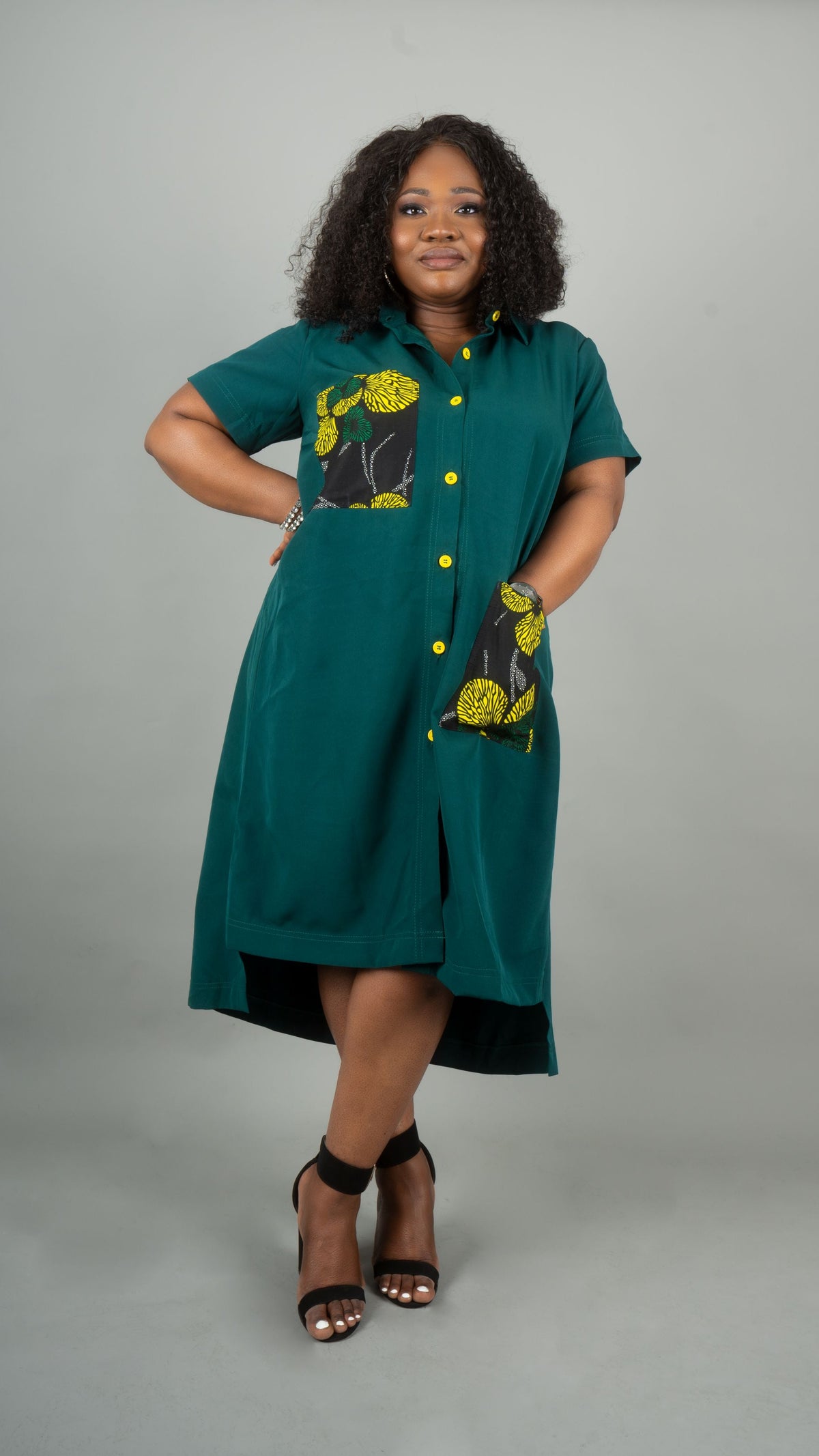 A woman modeling a teal green short-sleeve button-up dress with yellow and black floral patterned pockets, standing against a solid gray studio background at Bahati Design.