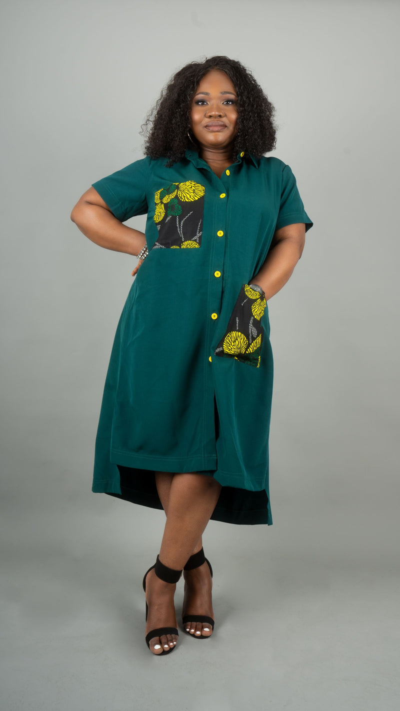 A woman modeling a teal green short-sleeve button-up dress with yellow and black floral patterned pockets, standing against a solid gray studio background at Bahati Design.