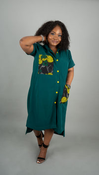A woman modeling a teal green short-sleeve button-up dress with yellow and black floral patterned pockets, standing against a solid gray studio background at Bahati Design.