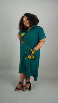 A woman modeling a short-sleeve teal green midi shirt dress with yellow and black patterned pockets, standing against a solid gray studio background at Bahati Design.