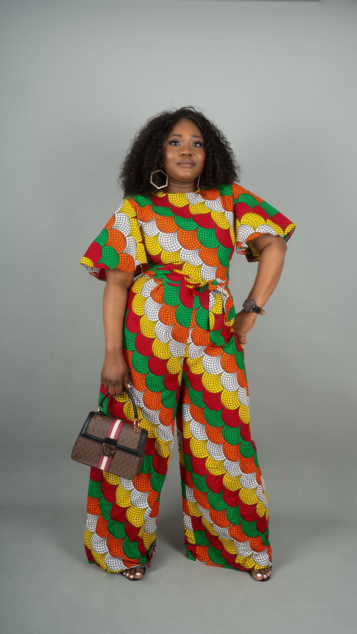 A woman modeling a matching two-piece set with a vibrant red, green, yellow, and white patterned fabric. The set includes a short-sleeve top with a tie waist and matching wide-leg pants at Bahati Design