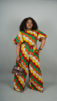 A woman modeling a matching two-piece set with a vibrant red, green, yellow, and white patterned fabric. The set includes a short-sleeve top with a tie waist and matching wide-leg pants at Bahati Design