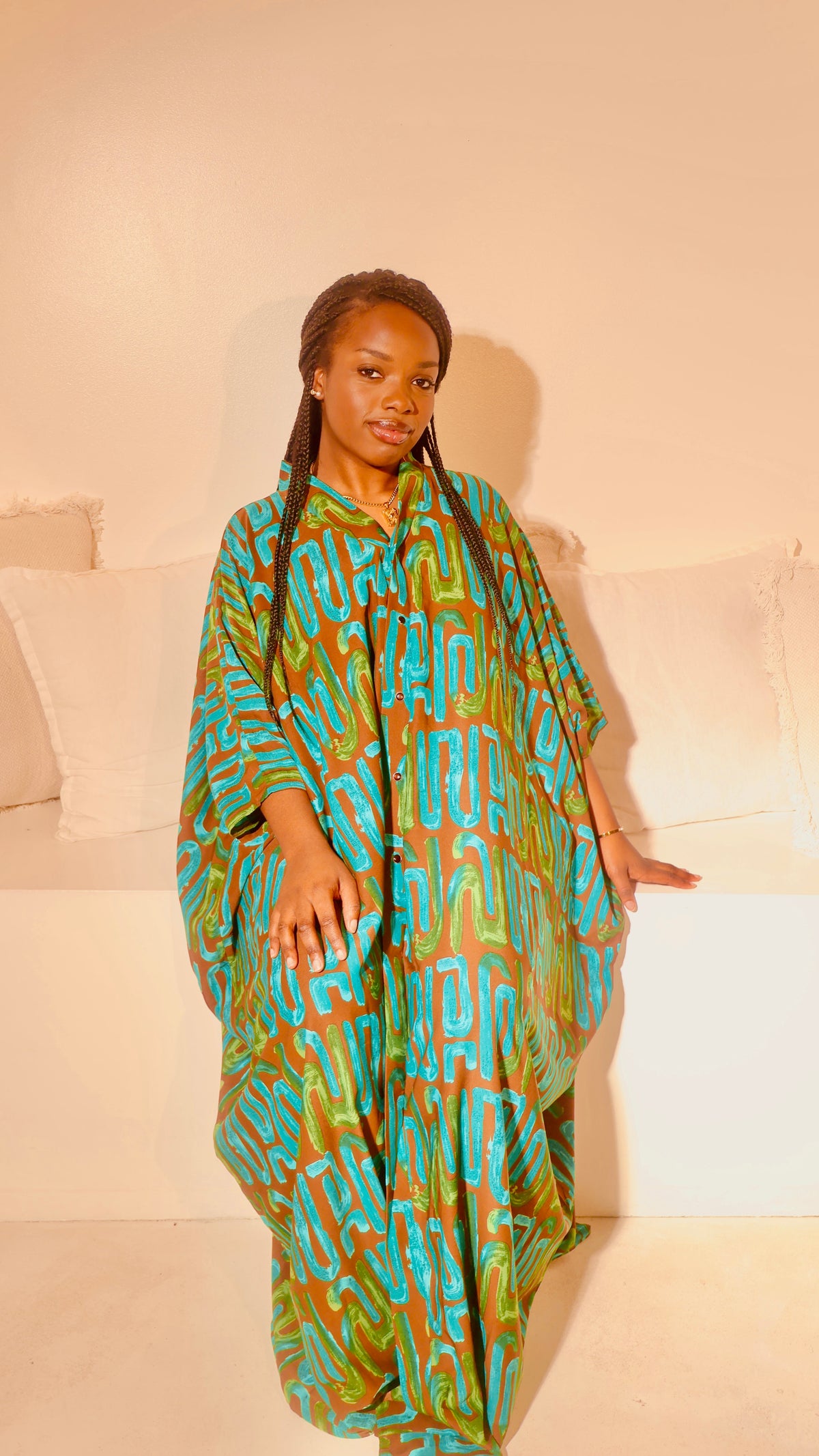 A woman wearing a loose, long kaftan dress with a vibrant turquoise and olive green abstract pattern, standing in front of a white bench with pillows at Bahait Design