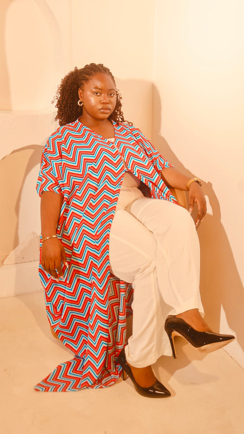A woman posing in a bright, warm-toned room wearing a short-sleeve chevron-patterned tunic top with red, blue, and white zigzags, paired with white pants and black heels.