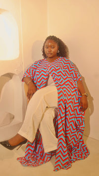 A woman sits on a white chair in a minimalist room, wearing a vibrant red, blue, and white chevron-patterned tunic draped over white wide-leg pants and black high heels.