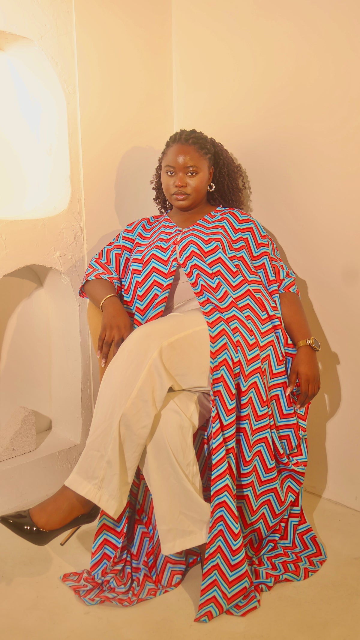 A woman sits on a white chair in a minimalist room, wearing a vibrant red, blue, and white chevron-patterned tunic draped over white wide-leg pants and black high heels.