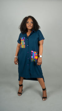 A woman modeling a dark teal button-down shirt dress with colorful geometric patterned pocket details, standing against a solid gray studio background at Bahati Design.