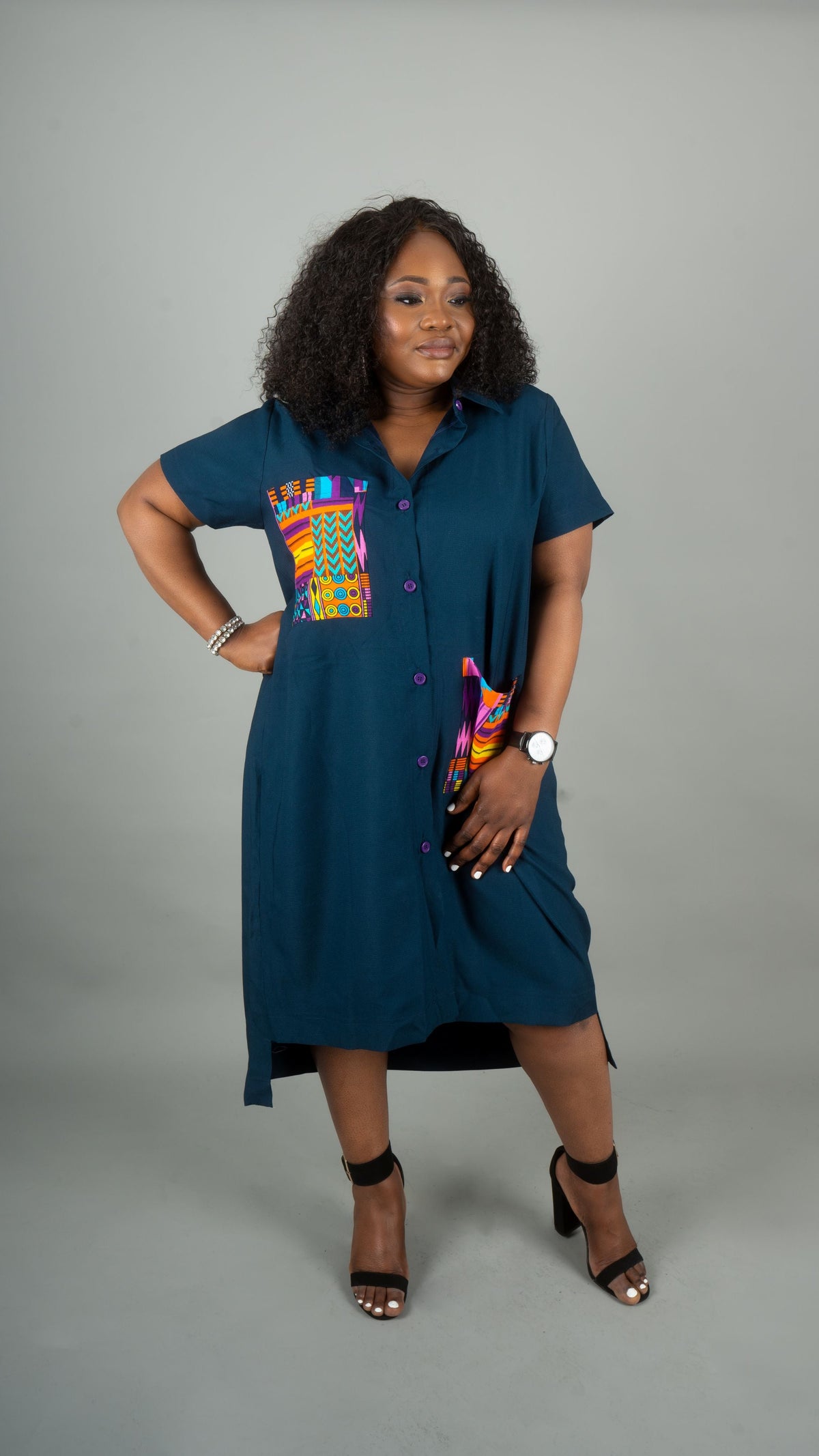 A woman modeling a short-sleeve button-front dress in deep teal blue with a colorful geometric patterned chest panel, standing against a plain gray studio background at Bahati Design.