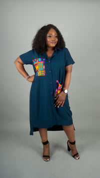 A woman modeling a short-sleeve button-front dress in deep teal blue with a colorful geometric patterned chest panel, standing against a plain gray studio background at Bahati Design.