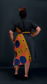 A woman is shown from behind wearing a black short-sleeve dress with a vibrant multicolor African wax print skirt panel and matching belt, set against a dark studio background.