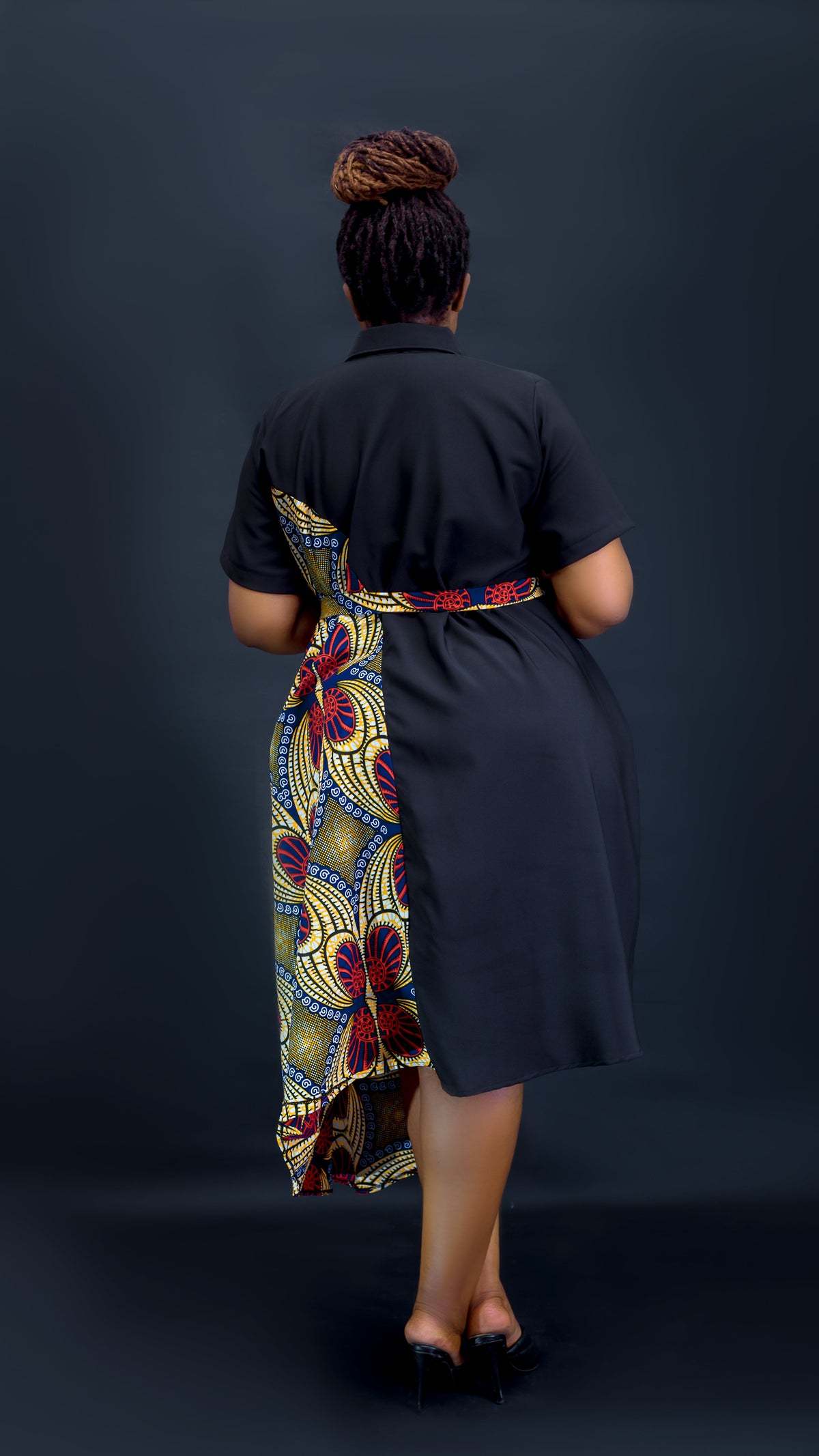 A woman viewed from behind wearing a black short-sleeve dress with a colorful patterned wrap skirt and waist tie, standing against a dark studio background at Bahai Design.