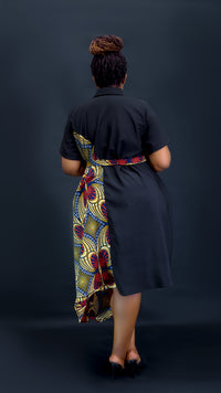 A woman viewed from behind wearing a black short-sleeve dress with a colorful patterned wrap skirt and waist tie, standing against a dark studio background at Bahai Design.