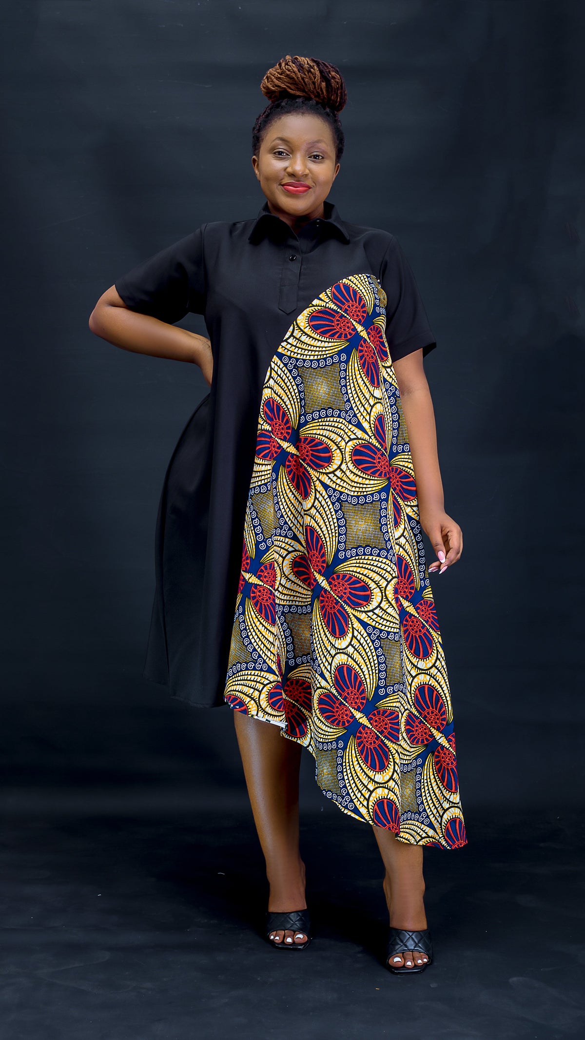 A woman modeling a short-sleeve dress with a black shirt-style overlay and an asymmetrical draped panel featuring a bold yellow, red, blue, and black geometric floral pattern, posed against a dark studio backdrop at Bahati Design