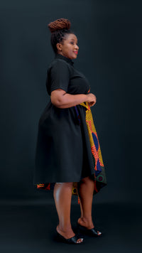 A woman stands in profile wearing a black short-sleeve dress with a colorful patterned fabric draped over one side, posed against a dark studio background.