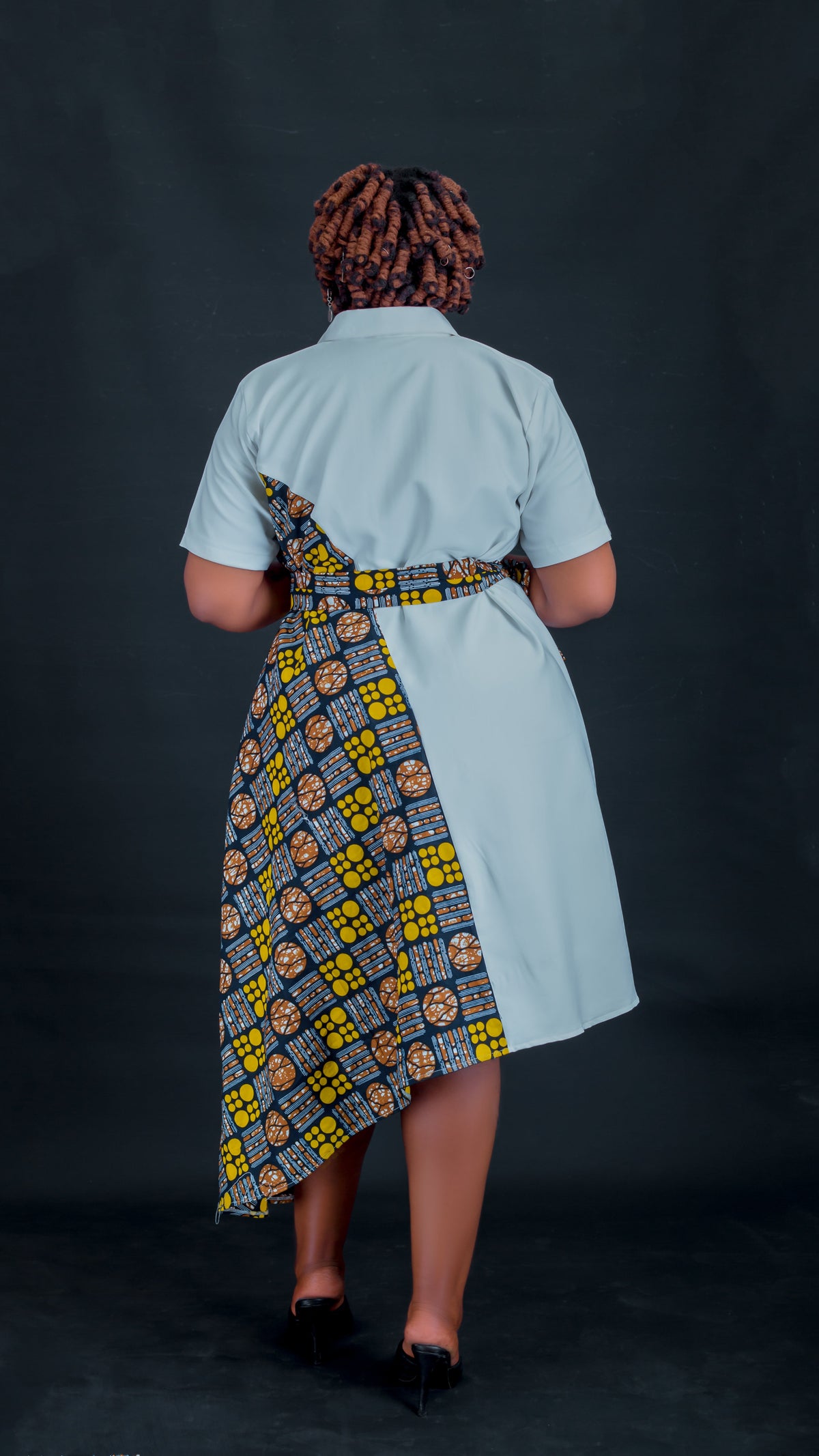 A woman viewed from behind wearing a short-sleeve light blue dress with a contrasting patterned waist panel in yellow, black, and blue, set against a dark studio background.