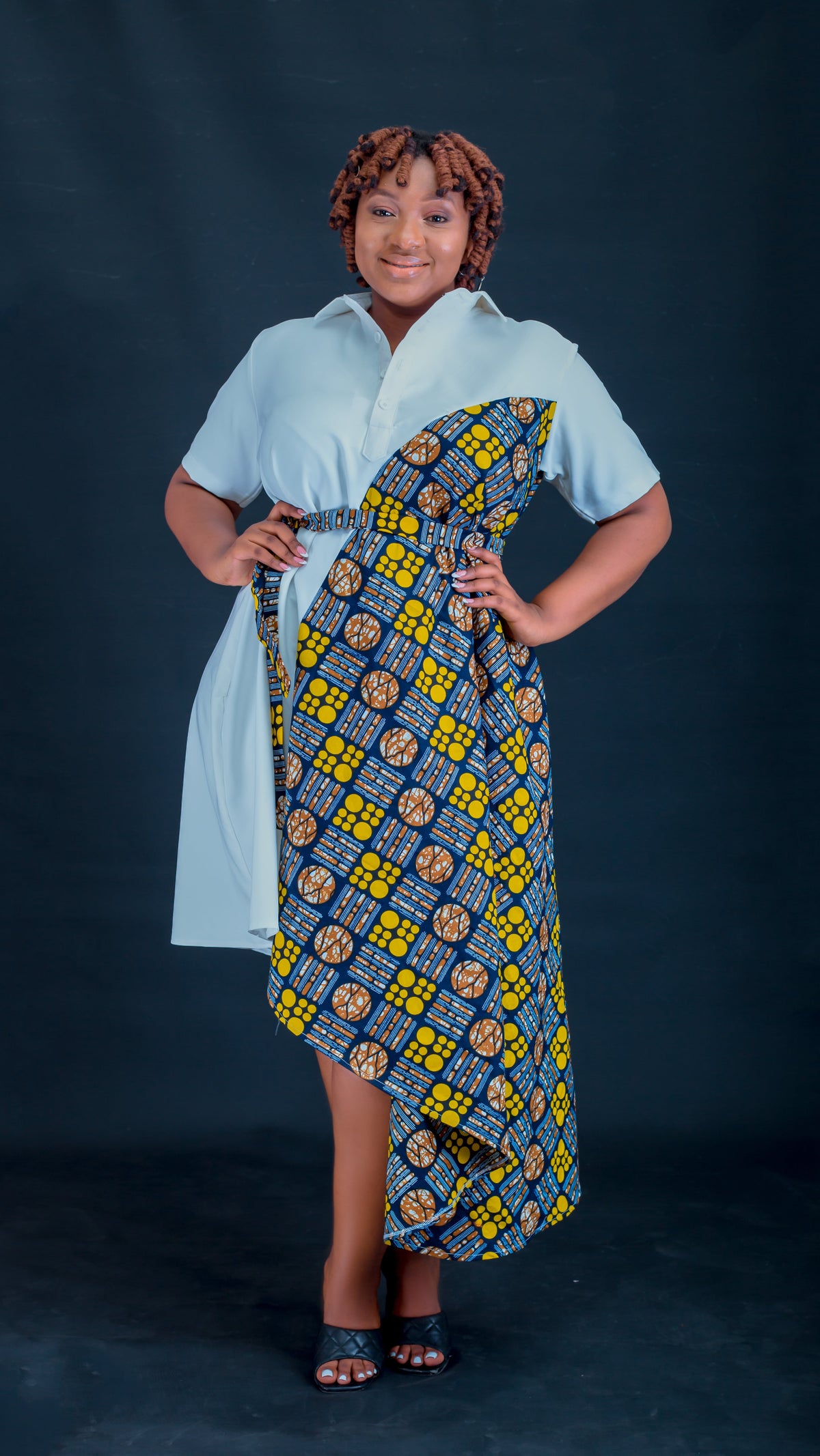 A woman modeling a short-sleeve dress with a light blue shirt-style overlay and a draped waist panel featuring a yellow, blue, and brown geometric African wax print, posed against a dark studio backdrop.