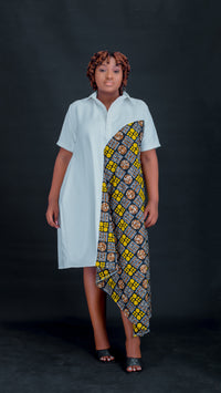 A woman modeling a short-sleeve collared dress with an asymmetrical design, featuring a solid light blue left side and a patterned yellow, black, and white African wax print panel on the right side, set against a dark studio background. Bahati Design