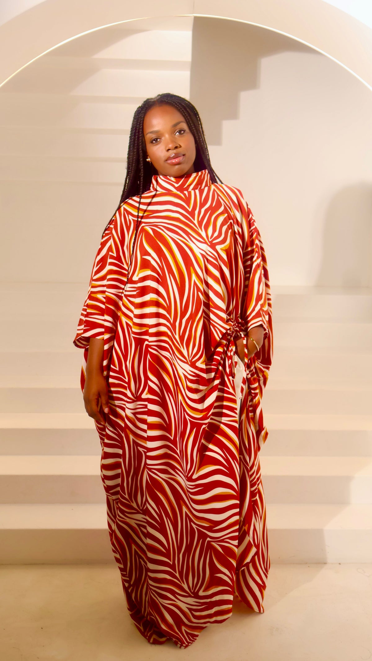 A woman wearing a high-neck, long-sleeved kaftan dress with a bold red and white zebra-like abstract pattern, standing in front of a minimalist white staircase at Bahati Design