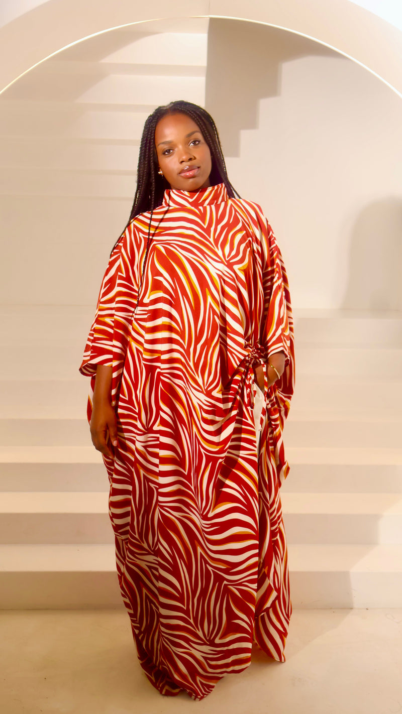 A woman wearing a high-neck, long-sleeved kaftan dress with a bold red and white zebra-like abstract pattern, standing in front of a minimalist white staircase at Bahati Design