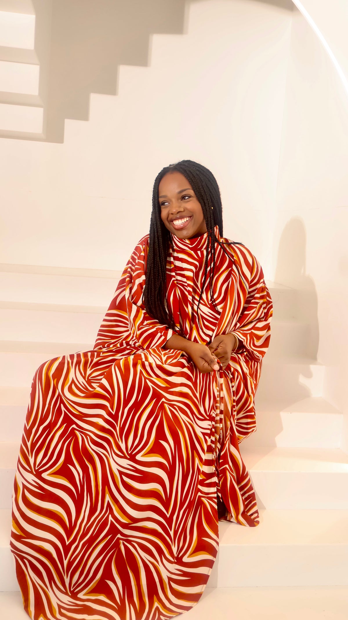A woman with long black braids sits smiling on white steps, wearing a flowing high-neck dress with a bold red, orange, and white wavy pattern.