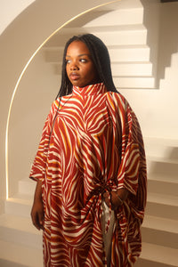 A woman with long black braids stands in front of white steps wearing a high-neck, loose-fitting kaftan dress with a bold red, orange, and cream wavy abstract pattern at Bahati Design.