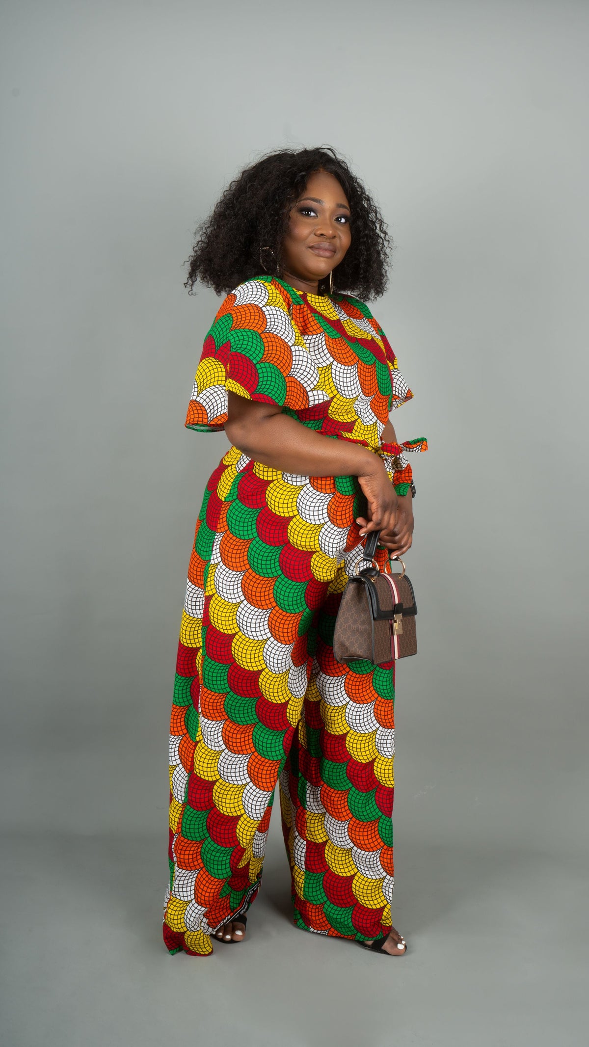 A woman modeling a matching two-piece set featuring a vibrant, multicolor African wax print with a scallop pattern in red, green, yellow, and white. The set includes a short-sleeve top and wide-leg trousers, styled with a small brown handbag against a solid gray studio background.