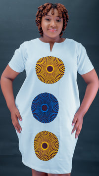 A woman modeling a white short-sleeve dress with three large circular African wax print motifs in yellow, blue, and red, set against a dark gray background.