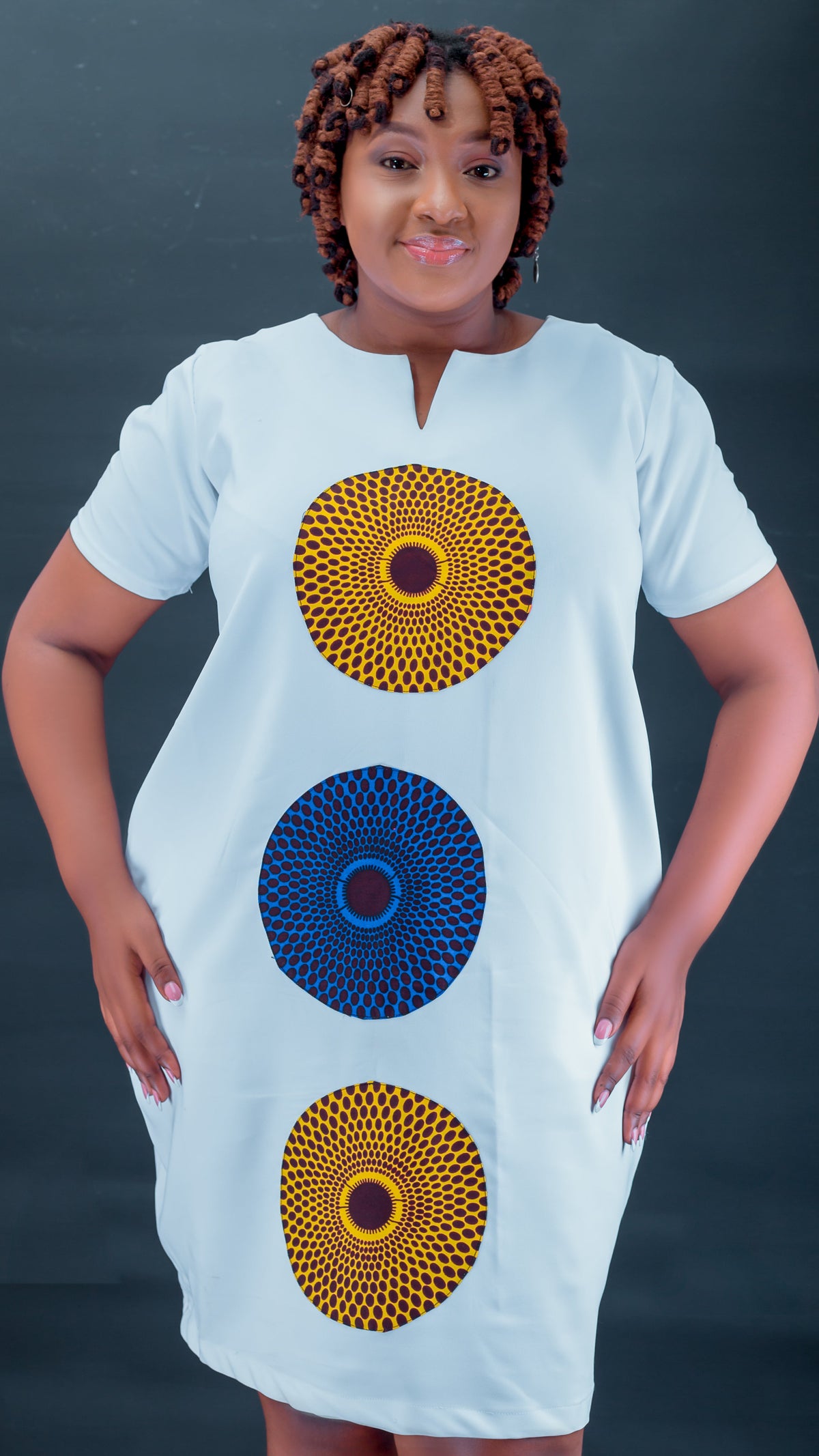 A woman modeling a white short-sleeve dress with three large circular African wax print motifs in yellow, blue, and red, set against a dark gray background.