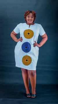 A woman modeling a white short-sleeve dress with three vertical circular patterns in yellow, blue, and yellow on the front, posed against a dark blue studio backdrop.
