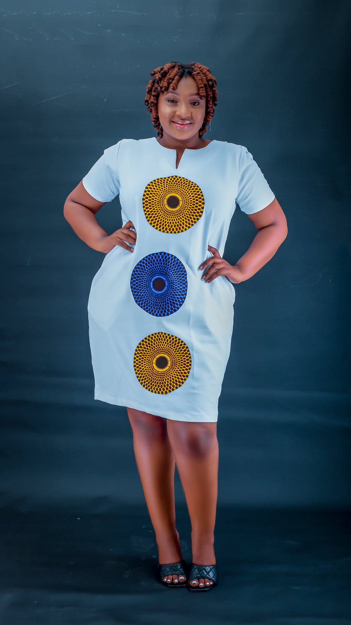 A woman modeling a white short-sleeve dress with three vertical circular patterns in yellow, blue, and yellow on the front, posed against a dark blue studio backdrop.