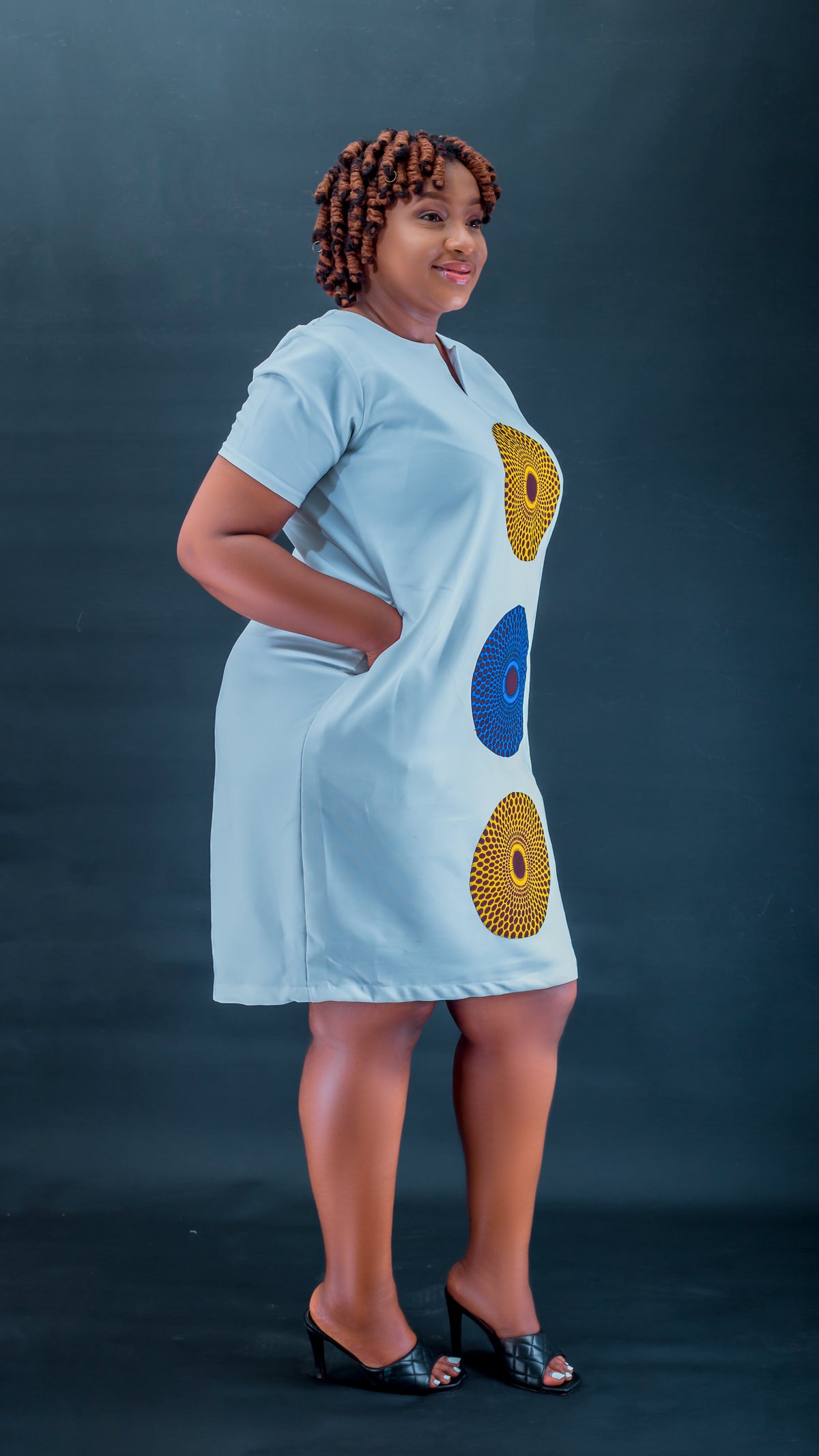 A woman modeling a light blue short-sleeve dress with three large circular African wax print motifs in yellow, blue, and red on the front, set against a dark blue studio background.