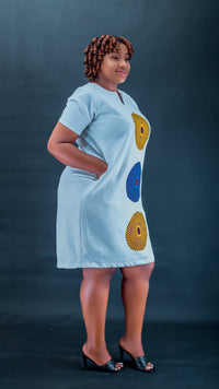 A woman modeling a light blue short-sleeve dress with three large circular African wax print motifs in yellow, blue, and red on the front, set against a dark blue studio background.