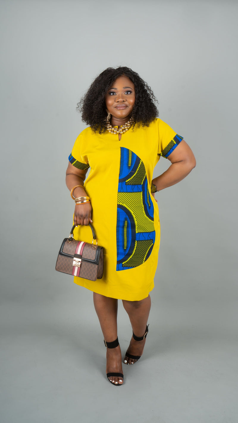 A woman modeling a bright yellow short-sleeve dress with blue and green geometric African wax print details, holding a brown handbag against a solid gray studio background.