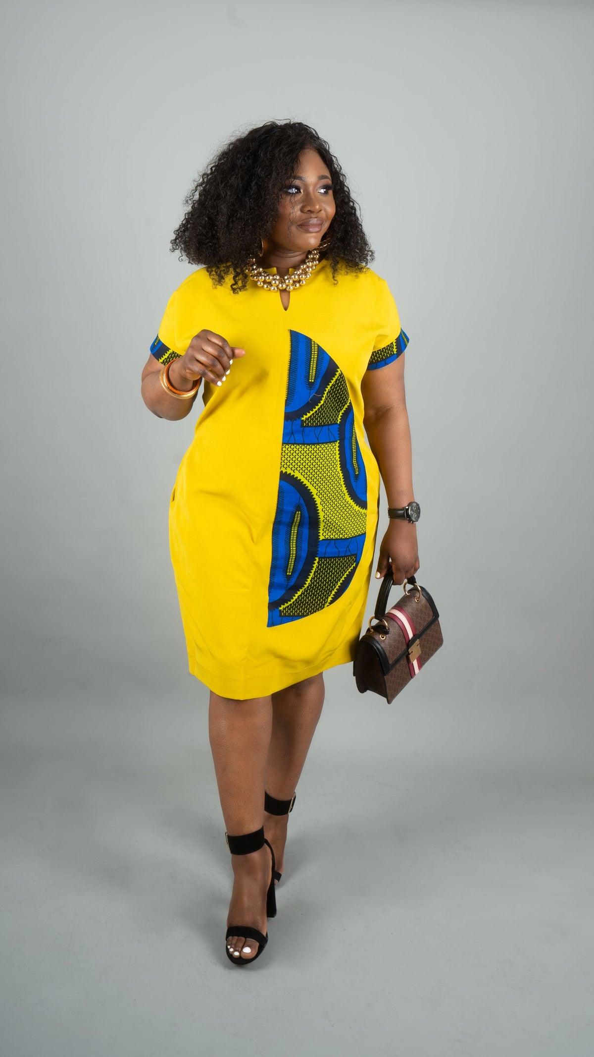A woman modeling a bright yellow short-sleeve dress with bold blue and green geometric patterns, styled with black strappy heels and holding a brown handbag against a solid gray studio background.