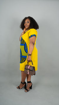 A woman modeling a bright yellow dress with bold blue and green geometric patterns, styled with black high heels and a brown handbag against a solid gray studio background.