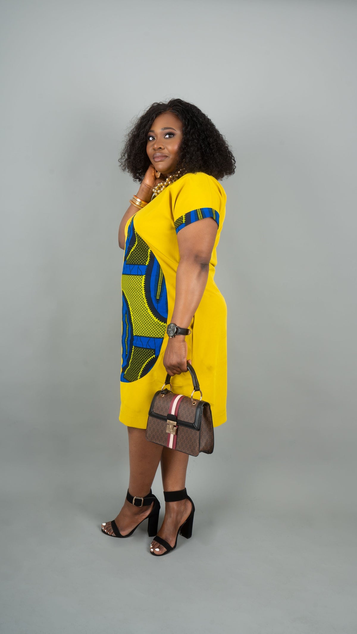 A woman modeling a bright yellow dress with bold blue and green geometric patterns, styled with black high heels and a brown handbag against a solid gray studio background.